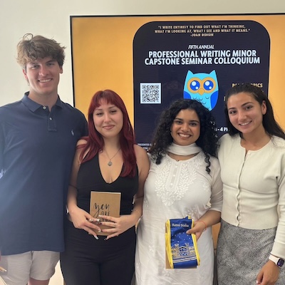Four students stand in front of a sign with a cartoon owl and the text "Fifth Annual Professional Writing Minor Capstone Seminar Colloquium"